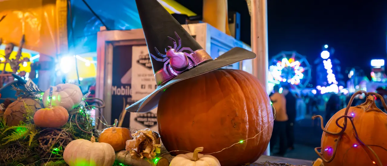 A pumpkin wearing a witch hat with a purple spider on its brim, string lights and mini pumpkins around it and the Ferris wheel glowing behind