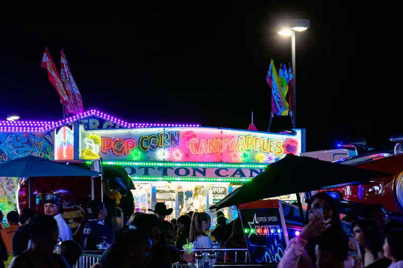 The neon popcorn, candy apple and cotton candy stand lit up at night with a crowd gathered in front of it