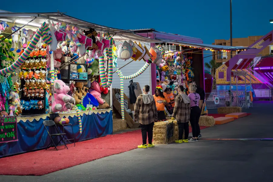 A midway game booth at dusk hung with plush prizes, friends playing at the counter