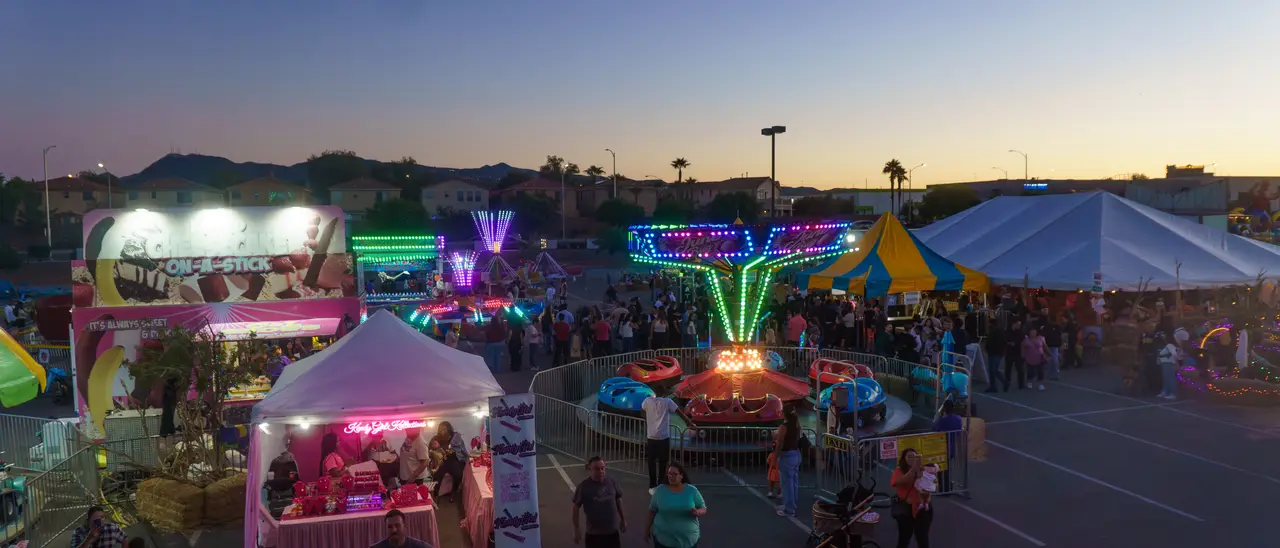 The midway at dusk from above: the swing ride and kiddie rides lit up, the big pumpkin tent, food stands and crowds of families walking between them