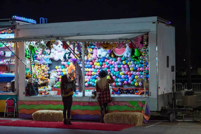 A guest at the balloon-pop trailer at night, its wall covered in balloons and plush prizes
