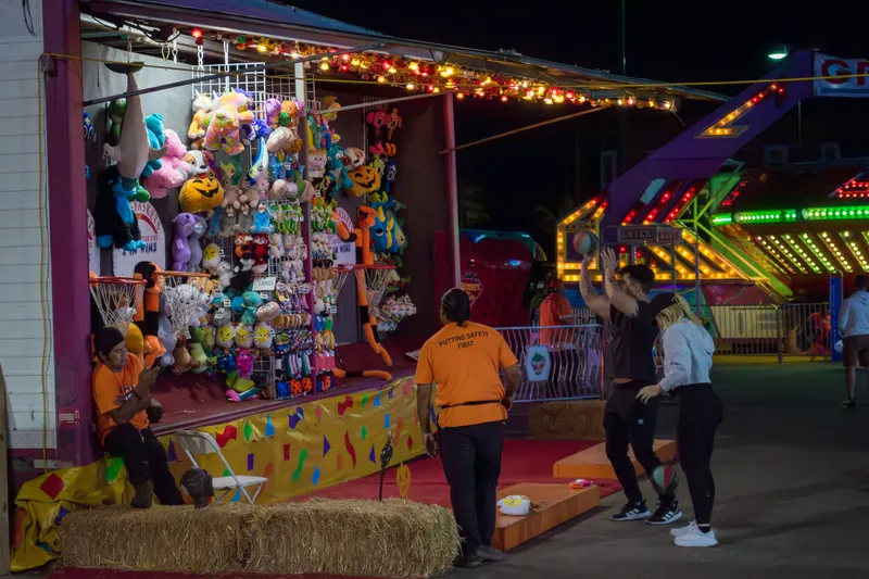 A guest shooting at a prize booth at night while staff watch, plush prizes hanging above and the rides lit behind