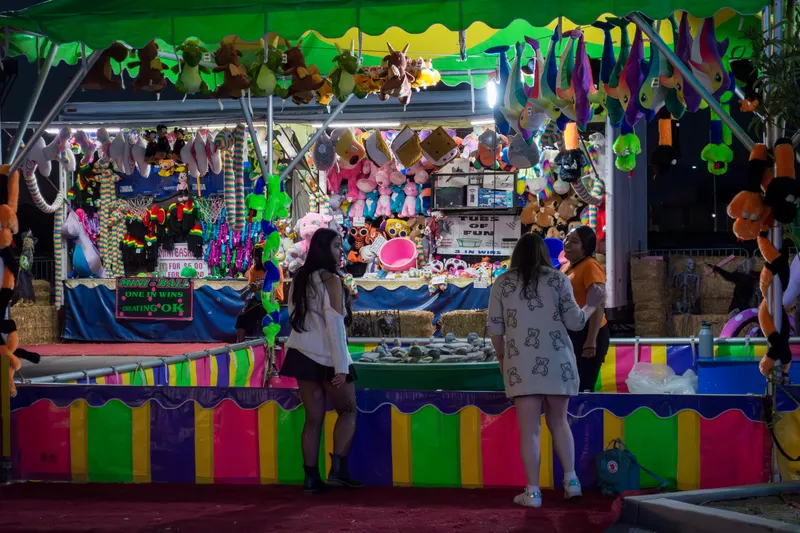 Two guests playing the Tubs of Fun toss at a rainbow-striped booth hung with plush prizes at night