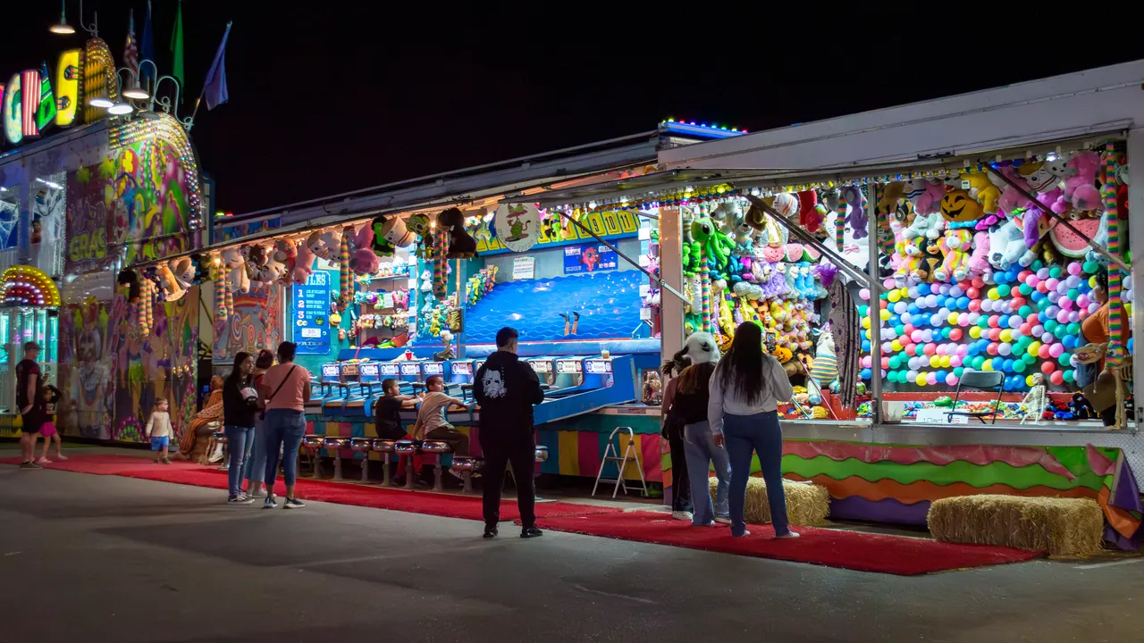 A row of lit game booths down the midway at night: the Water Gun Race with its line of seats, walls of plush prizes and the balloon-pop board, with guests playing at the counters and walking the red carpet