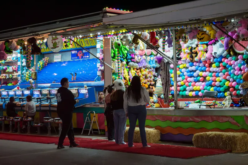 Guests playing the water race game at a midway booth at night, under a wall of plush prizes and a balloon-pop board