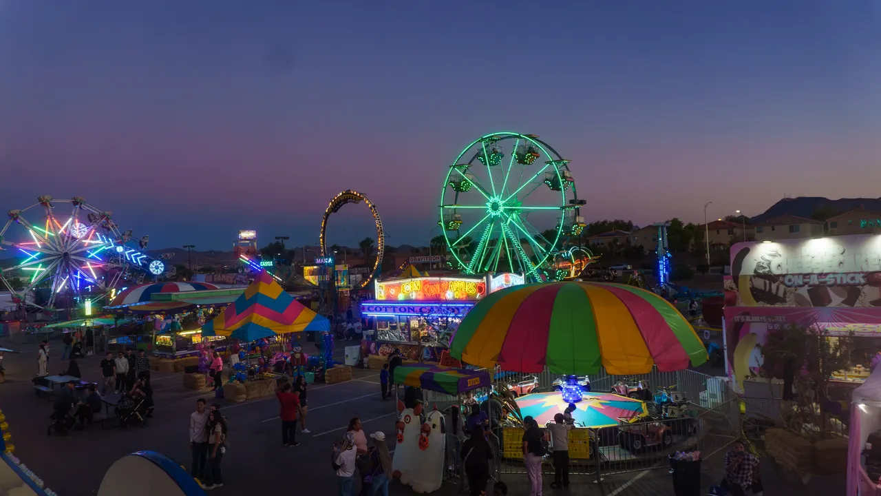 The Vegas Pumpkin Patch midway at dusk, seen from above: the rainbow-lit Ferris wheel, the Ring of Fire loop and the green Paradise Wheel over rows of game tents, the popcorn and cotton candy stand, the kiddie ride canopy and a crowd of families filling the midway