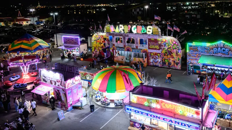 Vegas Pumpkin Patch at night from above: the Mardi Gras funhouse, the Water Gun Race and Toy Factory game booths, the popcorn and cotton candy stand, bumper cars and the kiddie canopy, with crowds in the midway and the Las Vegas skyline behind
